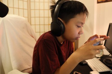 Güneydoğu Asyalı çocuk bilgisayar masasında akıllı telefonuyla oyun oynuyor..