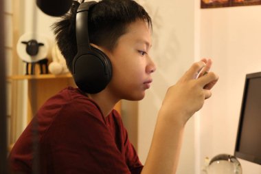 Güneydoğu Asyalı çocuk bilgisayar masasında akıllı telefonuyla oyun oynuyor..