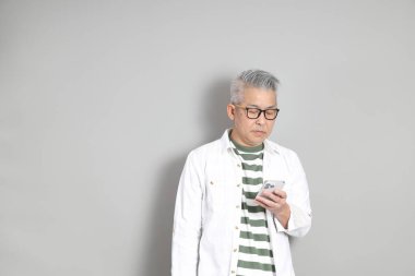 Doğu Asyalı bir adamın portresi gri saçlı ve gözlüklü, şık günlük kıyafetler giymiş, gri bir arka plana karşı duran.