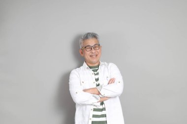 Doğu Asyalı bir adamın portresi gri saçlı ve gözlüklü, şık günlük kıyafetler giymiş, gri bir arka plana karşı duran.