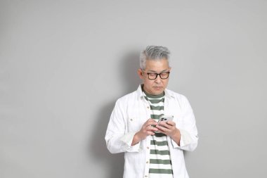 Doğu Asyalı bir adamın portresi gri saçlı ve gözlüklü, şık günlük kıyafetler giymiş, gri bir arka plana karşı duran.