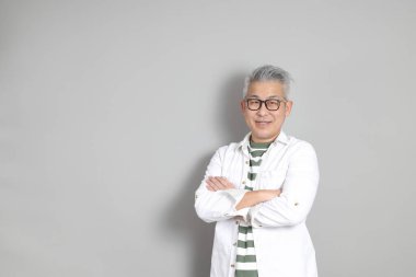 Doğu Asyalı bir adamın portresi gri saçlı ve gözlüklü, şık günlük kıyafetler giymiş, gri bir arka plana karşı duran.