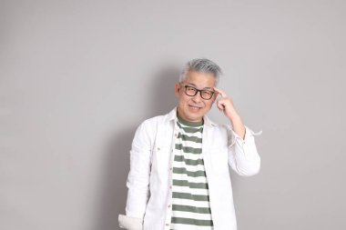 Doğu Asyalı bir adamın portresi gri saçlı ve gözlüklü, şık günlük kıyafetler giymiş, gri bir arka plana karşı duran.