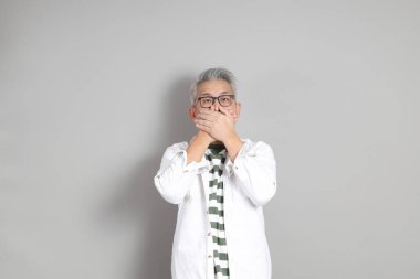 Doğu Asyalı bir adamın portresi gri saçlı ve gözlüklü, şık günlük kıyafetler giymiş, gri bir arka plana karşı duran.