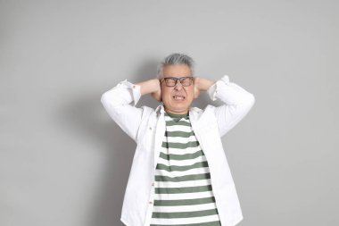 Doğu Asyalı bir adamın portresi gri saçlı ve gözlüklü, şık günlük kıyafetler giymiş, gri bir arka plana karşı duran.