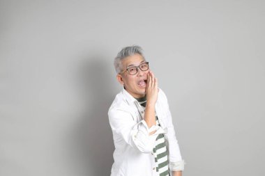 Doğu Asyalı bir adamın portresi gri saçlı ve gözlüklü, şık günlük kıyafetler giymiş, gri bir arka plana karşı duran.