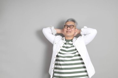 Doğu Asyalı bir adamın portresi gri saçlı ve gözlüklü, şık günlük kıyafetler giymiş, gri bir arka plana karşı duran.