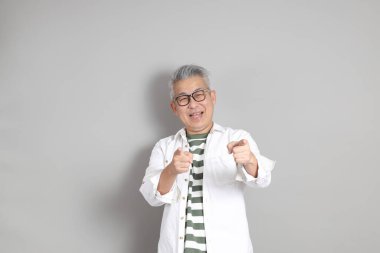 Doğu Asyalı bir adamın portresi gri saçlı ve gözlüklü, şık günlük kıyafetler giymiş, gri bir arka plana karşı duran.