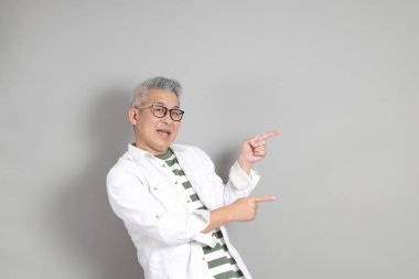 Doğu Asyalı bir adamın portresi gri saçlı ve gözlüklü, şık günlük kıyafetler giymiş, gri bir arka plana karşı duran.