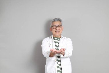 Doğu Asyalı bir adamın portresi gri saçlı ve gözlüklü, şık günlük kıyafetler giymiş, gri bir arka plana karşı duran.