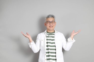 Doğu Asyalı bir adamın portresi gri saçlı ve gözlüklü, şık günlük kıyafetler giymiş, gri bir arka plana karşı duran.