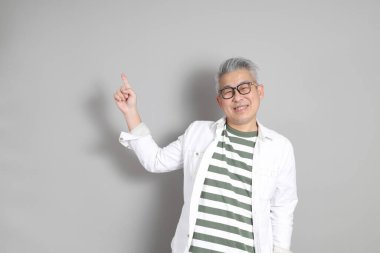 Doğu Asyalı bir adamın portresi gri saçlı ve gözlüklü, şık günlük kıyafetler giymiş, gri bir arka plana karşı duran.