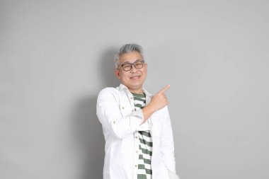 Doğu Asyalı bir adamın portresi gri saçlı ve gözlüklü, şık günlük kıyafetler giymiş, gri bir arka plana karşı duran.