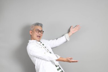 Doğu Asyalı bir adamın portresi gri saçlı ve gözlüklü, şık günlük kıyafetler giymiş, gri bir arka plana karşı duran.