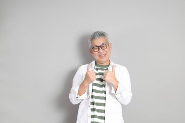 Doğu Asyalı bir adamın portresi gri saçlı ve gözlüklü, şık günlük kıyafetler giymiş, gri bir arka plana karşı duran.