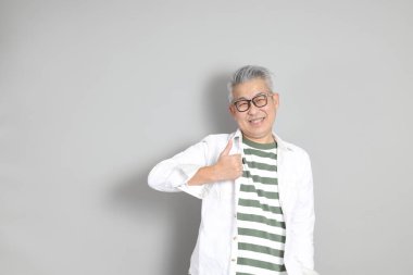 Doğu Asyalı bir adamın portresi gri saçlı ve gözlüklü, şık günlük kıyafetler giymiş, gri bir arka plana karşı duran.