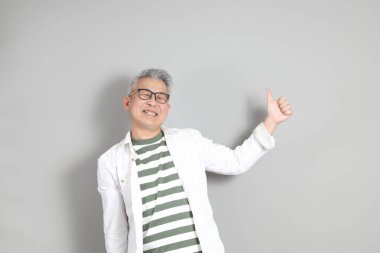 Doğu Asyalı bir adamın portresi gri saçlı ve gözlüklü, şık günlük kıyafetler giymiş, gri bir arka plana karşı duran.