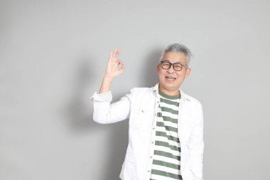 Doğu Asyalı bir adamın portresi gri saçlı ve gözlüklü, şık günlük kıyafetler giymiş, gri bir arka plana karşı duran.