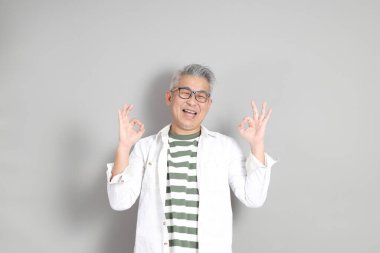 Doğu Asyalı bir adamın portresi gri saçlı ve gözlüklü, şık günlük kıyafetler giymiş, gri bir arka plana karşı duran.