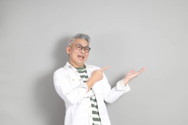 Doğu Asyalı bir adamın portresi gri saçlı ve gözlüklü, şık günlük kıyafetler giymiş, gri bir arka plana karşı duran.