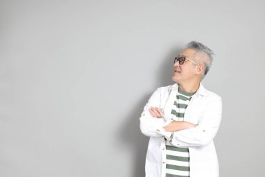 Doğu Asyalı bir adamın portresi gri saçlı ve gözlüklü, şık günlük kıyafetler giymiş, gri bir arka plana karşı duran.