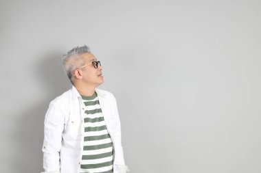 Doğu Asyalı bir adamın portresi gri saçlı ve gözlüklü, şık günlük kıyafetler giymiş, gri bir arka plana karşı duran.