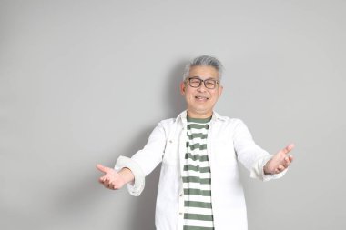 Doğu Asyalı bir adamın portresi gri saçlı ve gözlüklü, şık günlük kıyafetler giymiş, gri bir arka plana karşı duran.