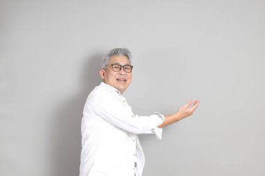 Doğu Asyalı bir adamın portresi gri saçlı ve gözlüklü, şık günlük kıyafetler giymiş, gri bir arka plana karşı duran.