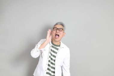 Doğu Asyalı bir adamın portresi gri saçlı ve gözlüklü, şık günlük kıyafetler giymiş, gri bir arka plana karşı duran.