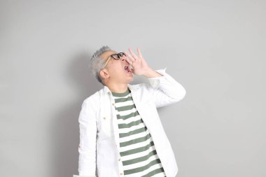 Doğu Asyalı bir adamın portresi gri saçlı ve gözlüklü, şık günlük kıyafetler giymiş, gri bir arka plana karşı duran.