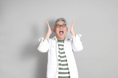 Doğu Asyalı bir adamın portresi gri saçlı ve gözlüklü, şık günlük kıyafetler giymiş, gri bir arka plana karşı duran.