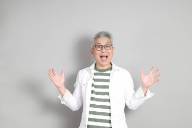 Doğu Asyalı bir adamın portresi gri saçlı ve gözlüklü, şık günlük kıyafetler giymiş, gri bir arka plana karşı duran.