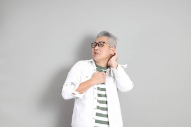 Doğu Asyalı bir adamın portresi gri saçlı ve gözlüklü, şık günlük kıyafetler giymiş, gri bir arka plana karşı duran.