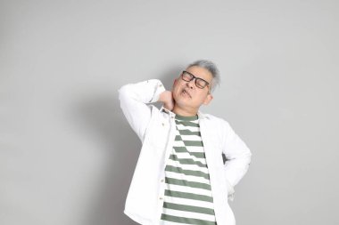 Doğu Asyalı bir adamın portresi gri saçlı ve gözlüklü, şık günlük kıyafetler giymiş, gri bir arka plana karşı duran.