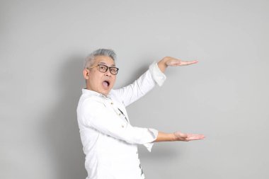 Doğu Asyalı bir adamın portresi gri saçlı ve gözlüklü, şık günlük kıyafetler giymiş, gri bir arka plana karşı duran.