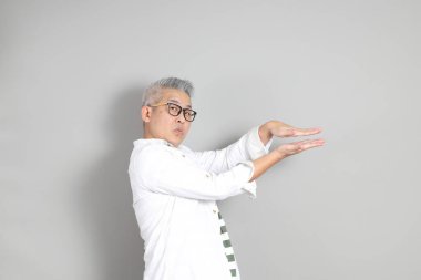 Doğu Asyalı bir adamın portresi gri saçlı ve gözlüklü, şık günlük kıyafetler giymiş, gri bir arka plana karşı duran.