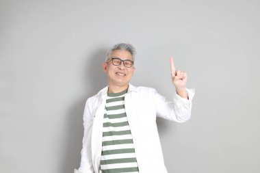 Doğu Asyalı bir adamın portresi gri saçlı ve gözlüklü, şık günlük kıyafetler giymiş, gri bir arka plana karşı duran.