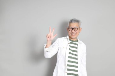 Doğu Asyalı bir adamın portresi gri saçlı ve gözlüklü, şık günlük kıyafetler giymiş, gri bir arka plana karşı duran.