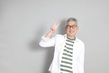 Doğu Asyalı bir adamın portresi gri saçlı ve gözlüklü, şık günlük kıyafetler giymiş, gri bir arka plana karşı duran.