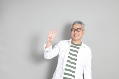 Doğu Asyalı bir adamın portresi gri saçlı ve gözlüklü, şık günlük kıyafetler giymiş, gri bir arka plana karşı duran.