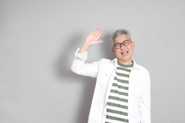 Doğu Asyalı bir adamın portresi gri saçlı ve gözlüklü, şık günlük kıyafetler giymiş, gri bir arka plana karşı duran.