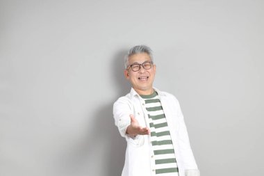 Doğu Asyalı bir adamın portresi gri saçlı ve gözlüklü, şık günlük kıyafetler giymiş, gri bir arka plana karşı duran.