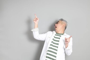 Doğu Asyalı bir adamın portresi gri saçlı ve gözlüklü, şık günlük kıyafetler giymiş, gri bir arka plana karşı duran.