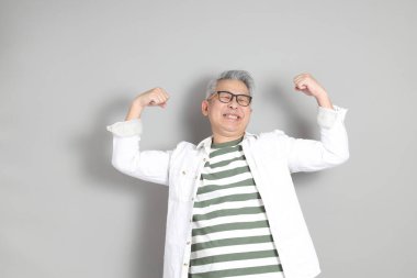 Doğu Asyalı bir adamın portresi gri saçlı ve gözlüklü, şık günlük kıyafetler giymiş, gri bir arka plana karşı duran.