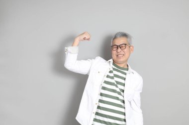 Doğu Asyalı bir adamın portresi gri saçlı ve gözlüklü, şık günlük kıyafetler giymiş, gri bir arka plana karşı duran.