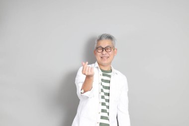 Doğu Asyalı bir adamın portresi gri saçlı ve gözlüklü, şık günlük kıyafetler giymiş, gri bir arka plana karşı duran.