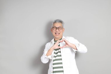 Doğu Asyalı bir adamın portresi gri saçlı ve gözlüklü, şık günlük kıyafetler giymiş, gri bir arka plana karşı duran.