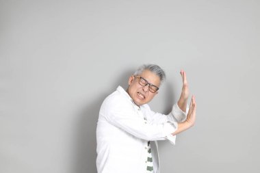 Doğu Asyalı bir adamın portresi gri saçlı ve gözlüklü, şık günlük kıyafetler giymiş, gri bir arka plana karşı duran.