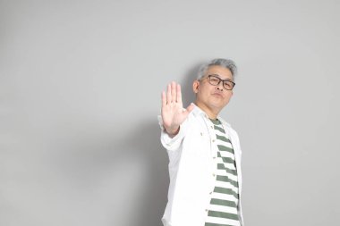 Doğu Asyalı bir adamın portresi gri saçlı ve gözlüklü, şık günlük kıyafetler giymiş, gri bir arka plana karşı duran.