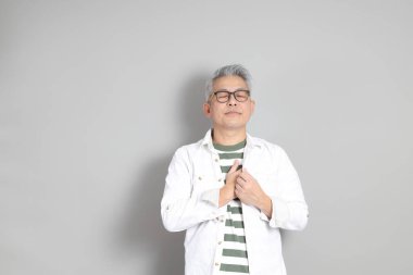 Doğu Asyalı bir adamın portresi gri saçlı ve gözlüklü, şık günlük kıyafetler giymiş, gri bir arka plana karşı duran.